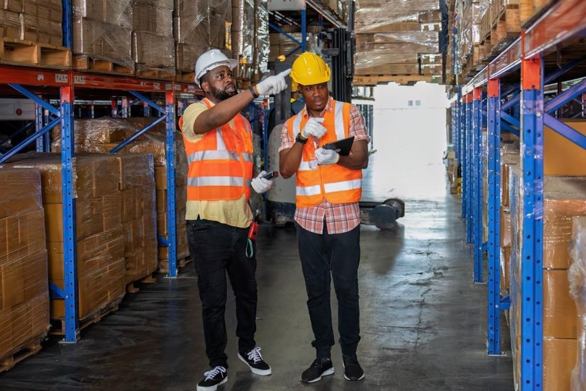 two warehouse workers performing a safety audit while wearing reflective vests and helmets
